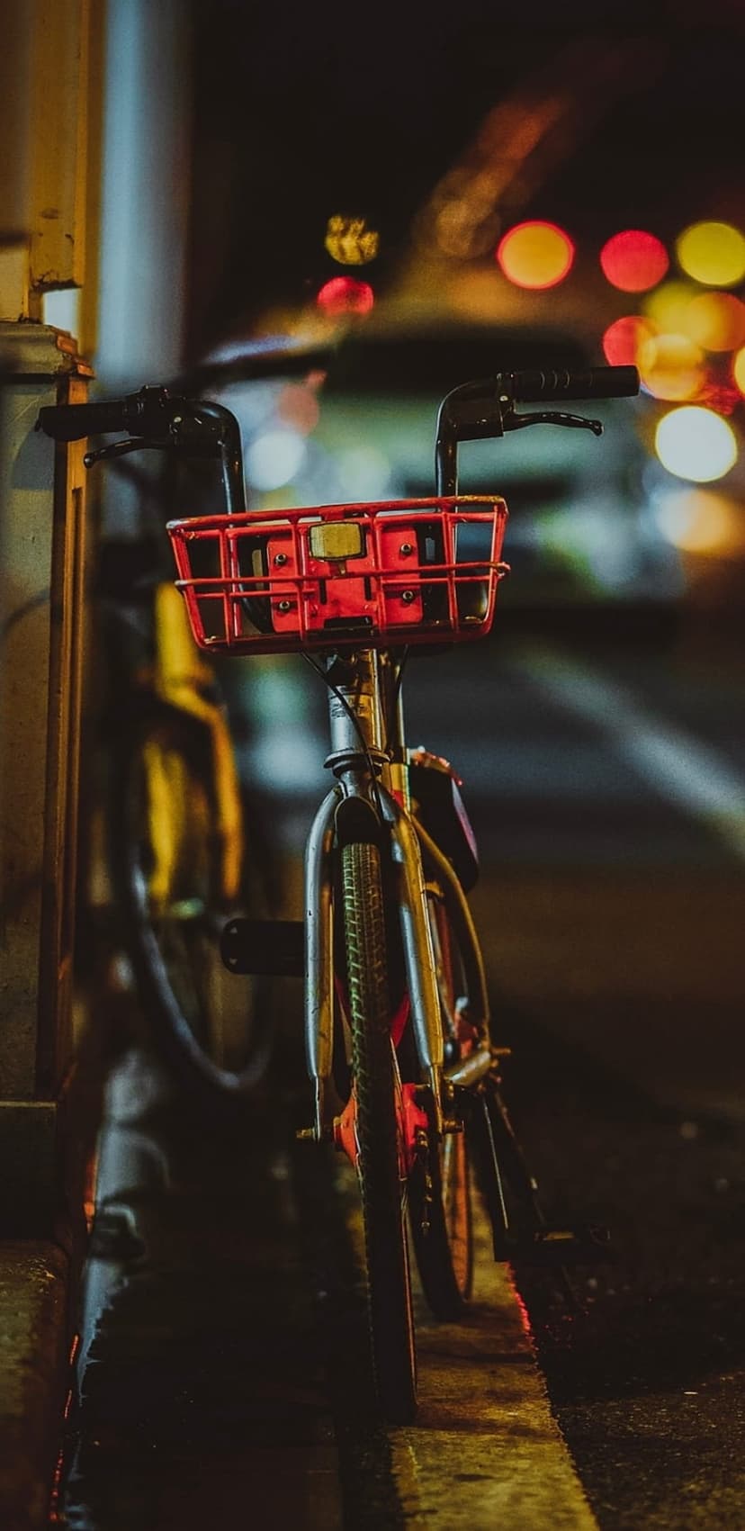 Red bicycle basket on chrome handlebars against a blurry background of golden and blue urban night bokeh lights.