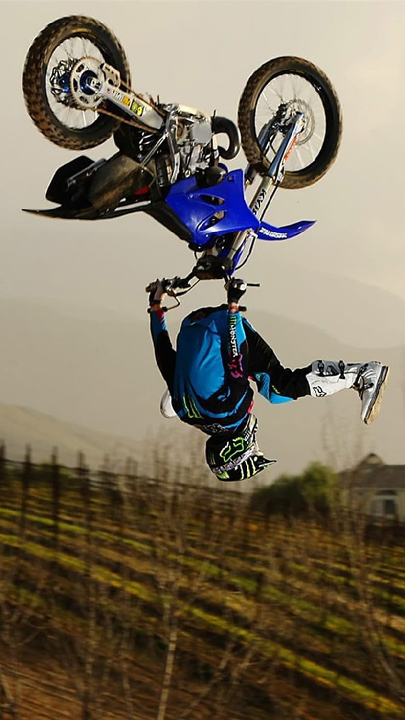 Motocross rider performing an inverted flip mid-air on a dirt bike against a hazy sky and blurred vineyard.