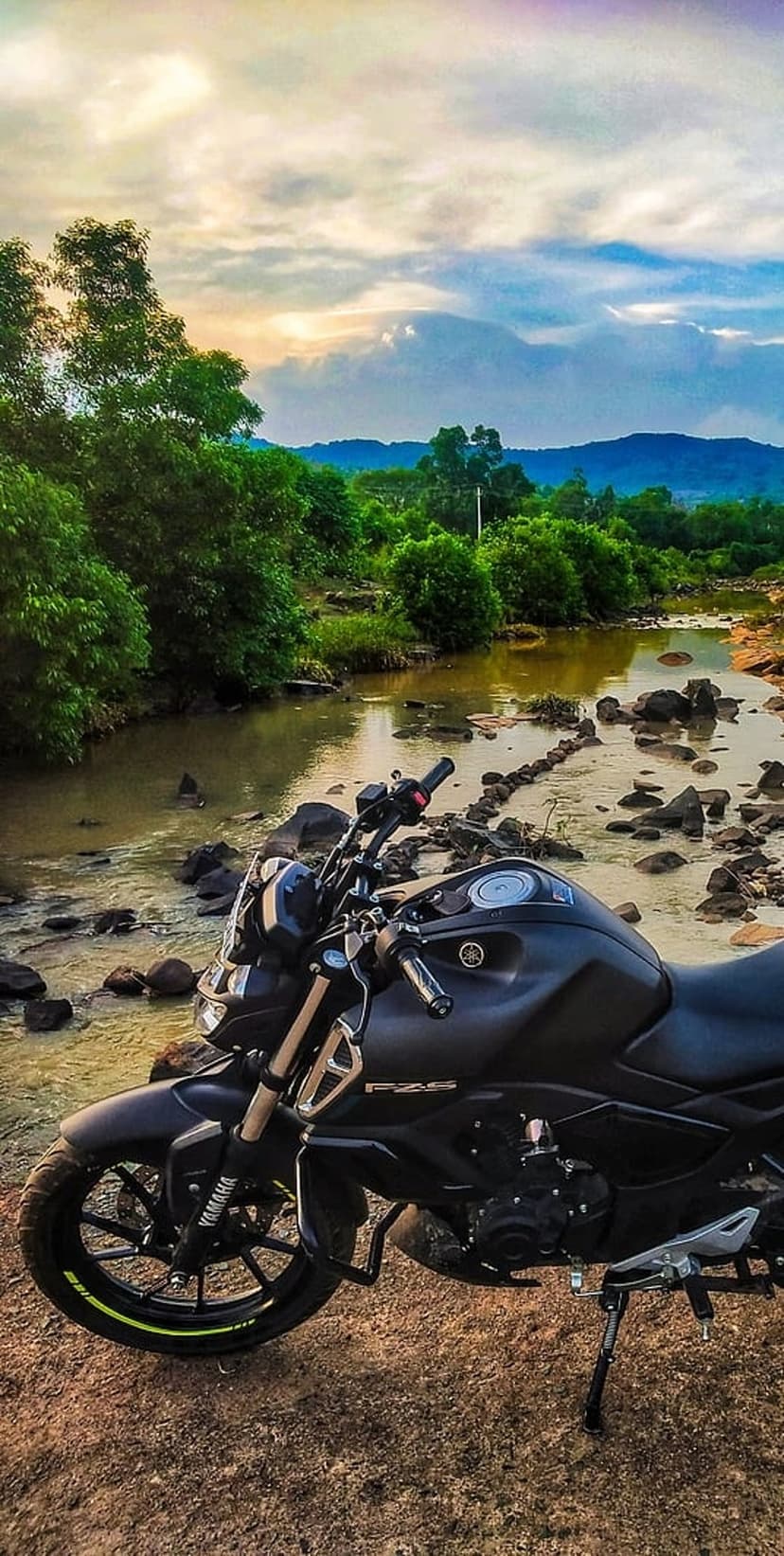 Black cruiser motorcycle parked on a rocky riverbank under a vibrant orange and purple sunset sky.