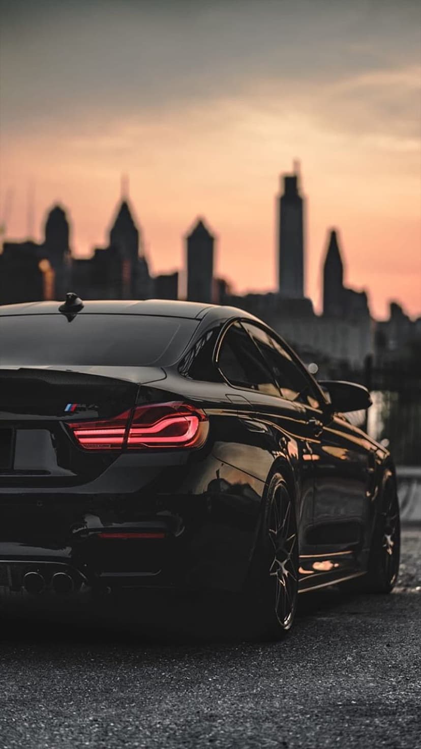 Black BMW M4 rear silhouette with glowing red taillights against a sunset urban skyline during golden hour.