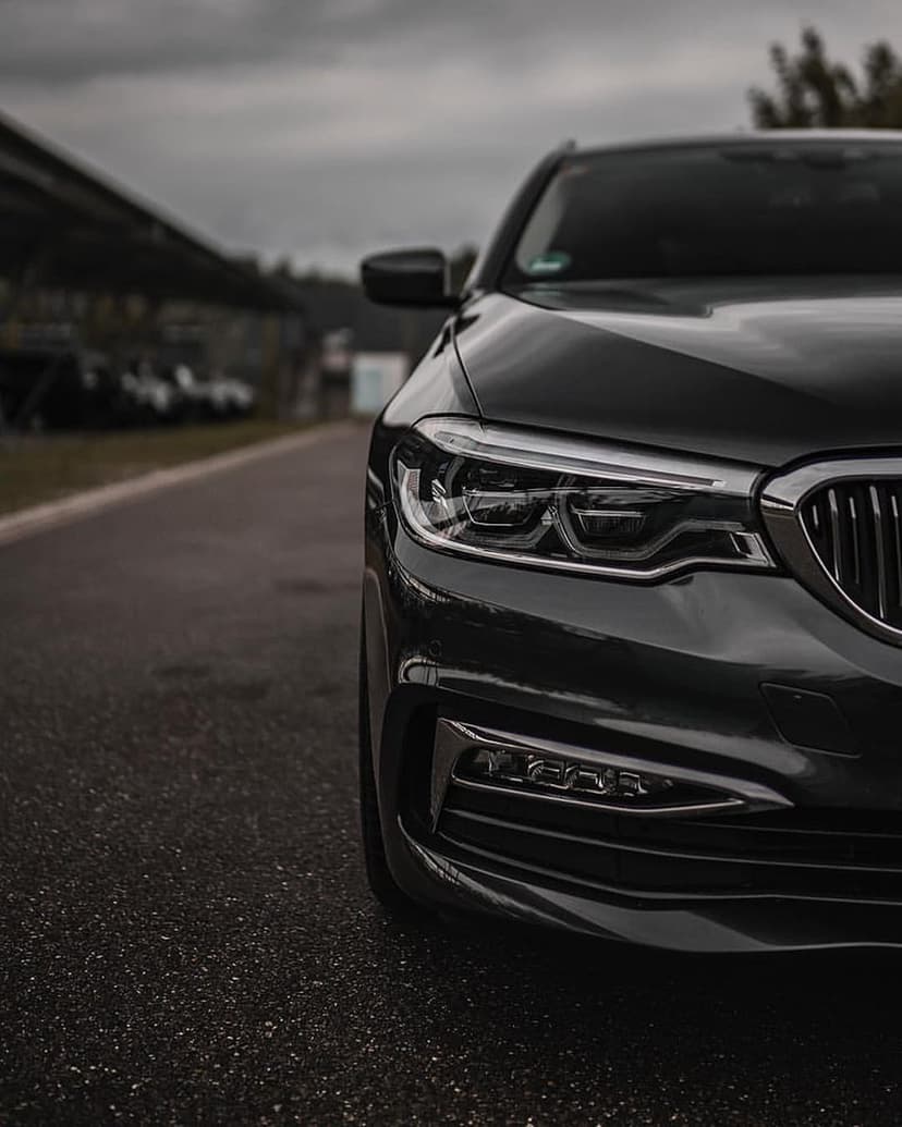 Black luxury sedan front grille and glowing LED headlights reflecting on a wet asphalt road under a moody sky.
