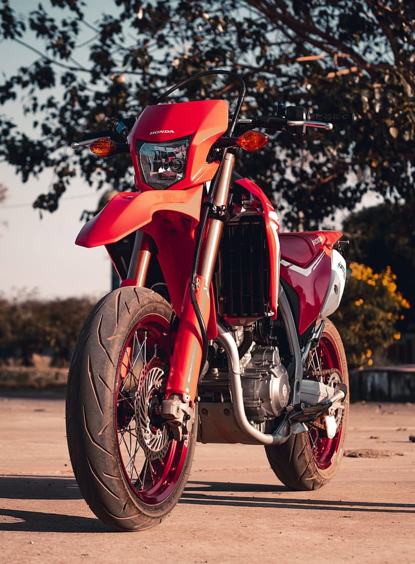 Honda supermoto motorcycle with red fairings and spoked wheels parked on a dirt path with blurred green foliage.