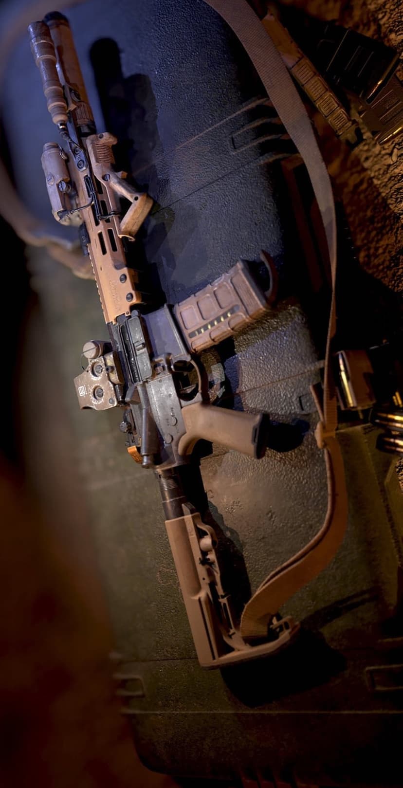 Tan tactical rifle with red dot sight and foregrip lying on a dark textured case next to brass ammunition.