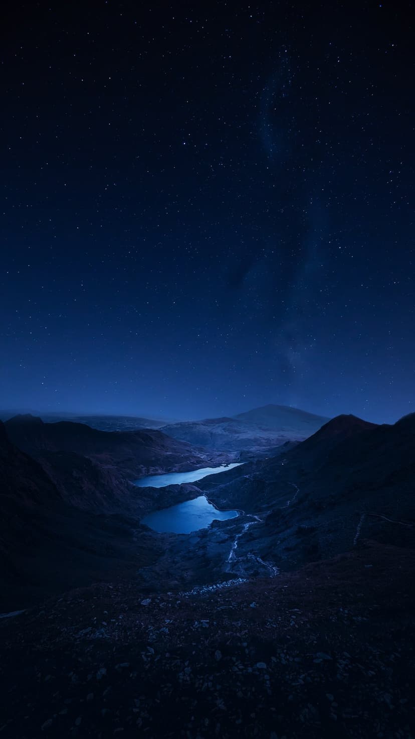 Milky Way stars shine over jagged mountain peaks and two glowing blue lakes connected by a stream in the valley.