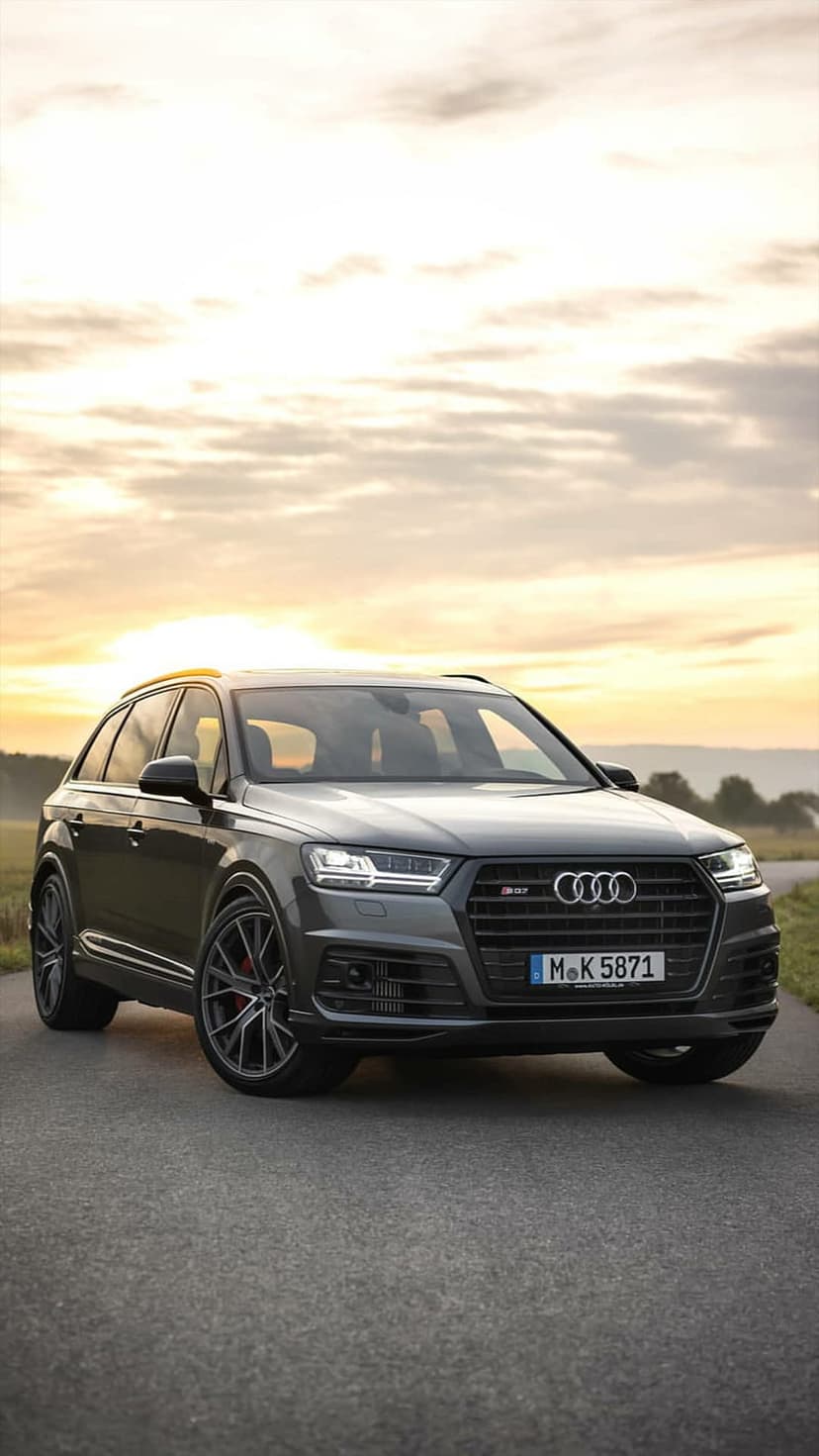 Dark grey Audi RS Q7 SUV with bright headlights parked on a paved road under a cloudy orange sunset sky.
