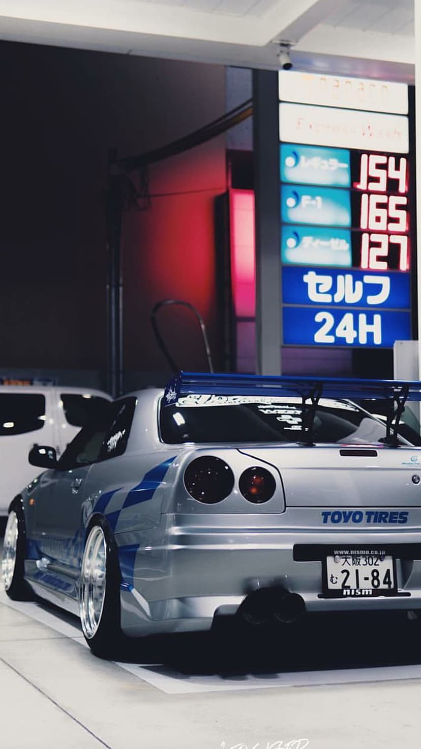 Silver Nissan Skyline GT-R R34 with blue stripes parked at a neon-lit Japanese gas station at night.