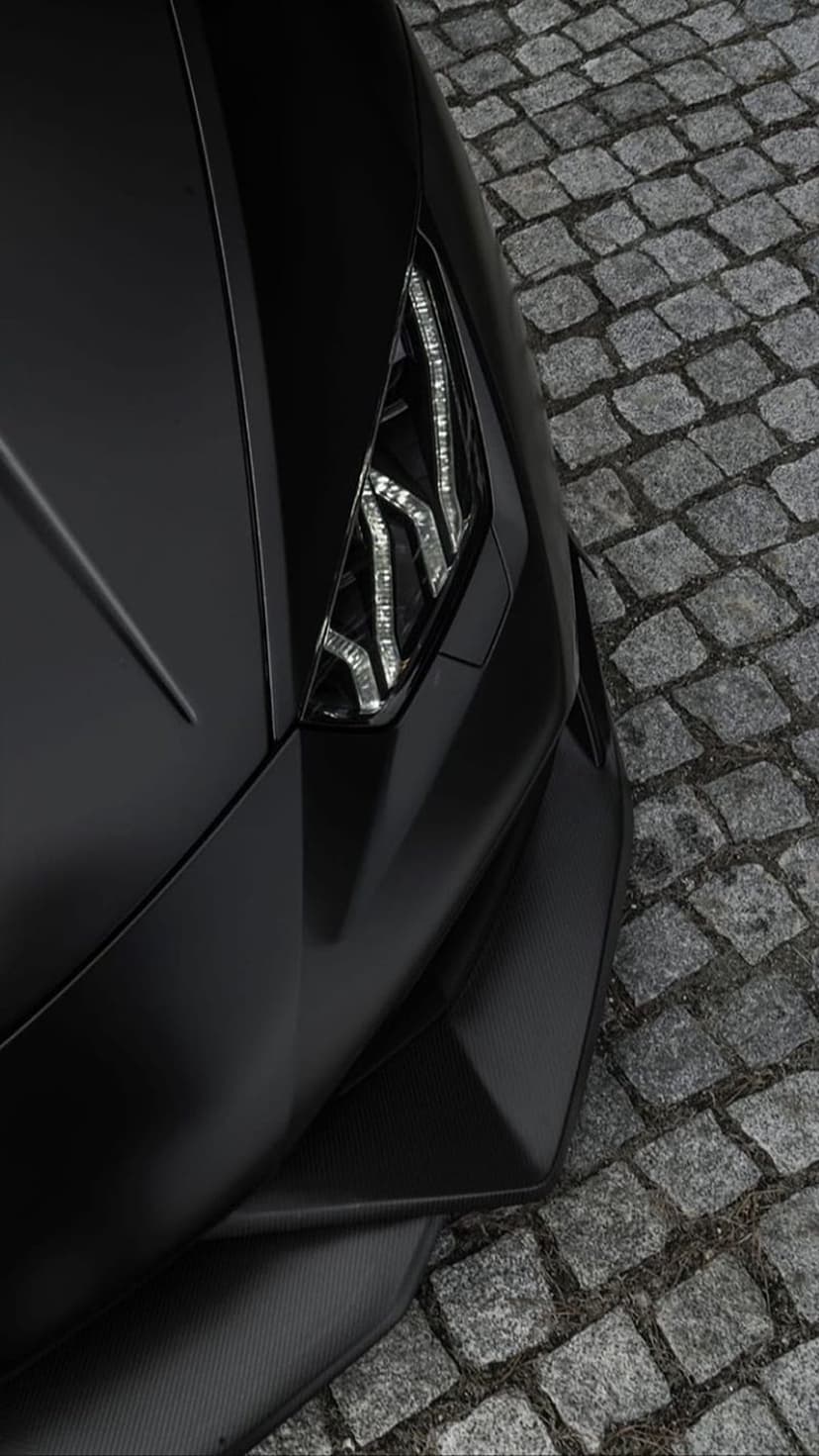 Matte black supercar headlight with glowing LED accents rests above a textured grey cobblestone road.