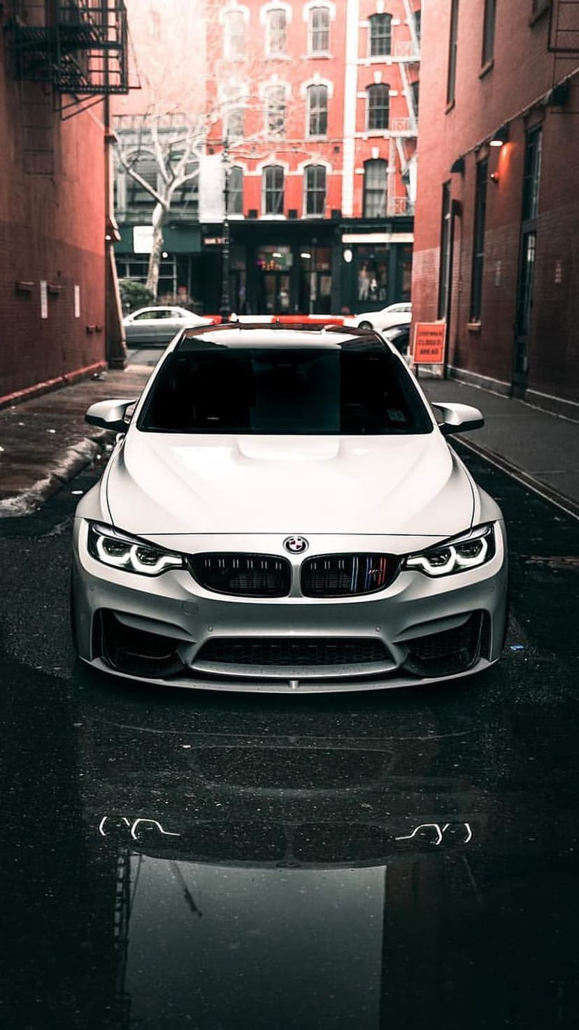 White BMW M4 sportscar parked on wet asphalt between old red brick buildings featuring iron fire escapes.