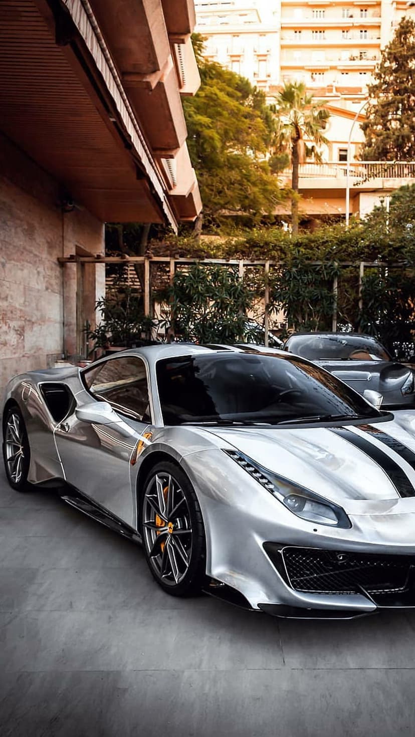 Silver Ferrari sports car with black racing stripes parked on a concrete path near lush greenery and glass walls.