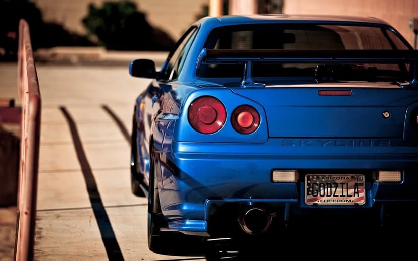 Rear of a blue Nissan Skyline GT-R R34 with round red taillights and a Godzilla Arizona license plate on a sunlit road.