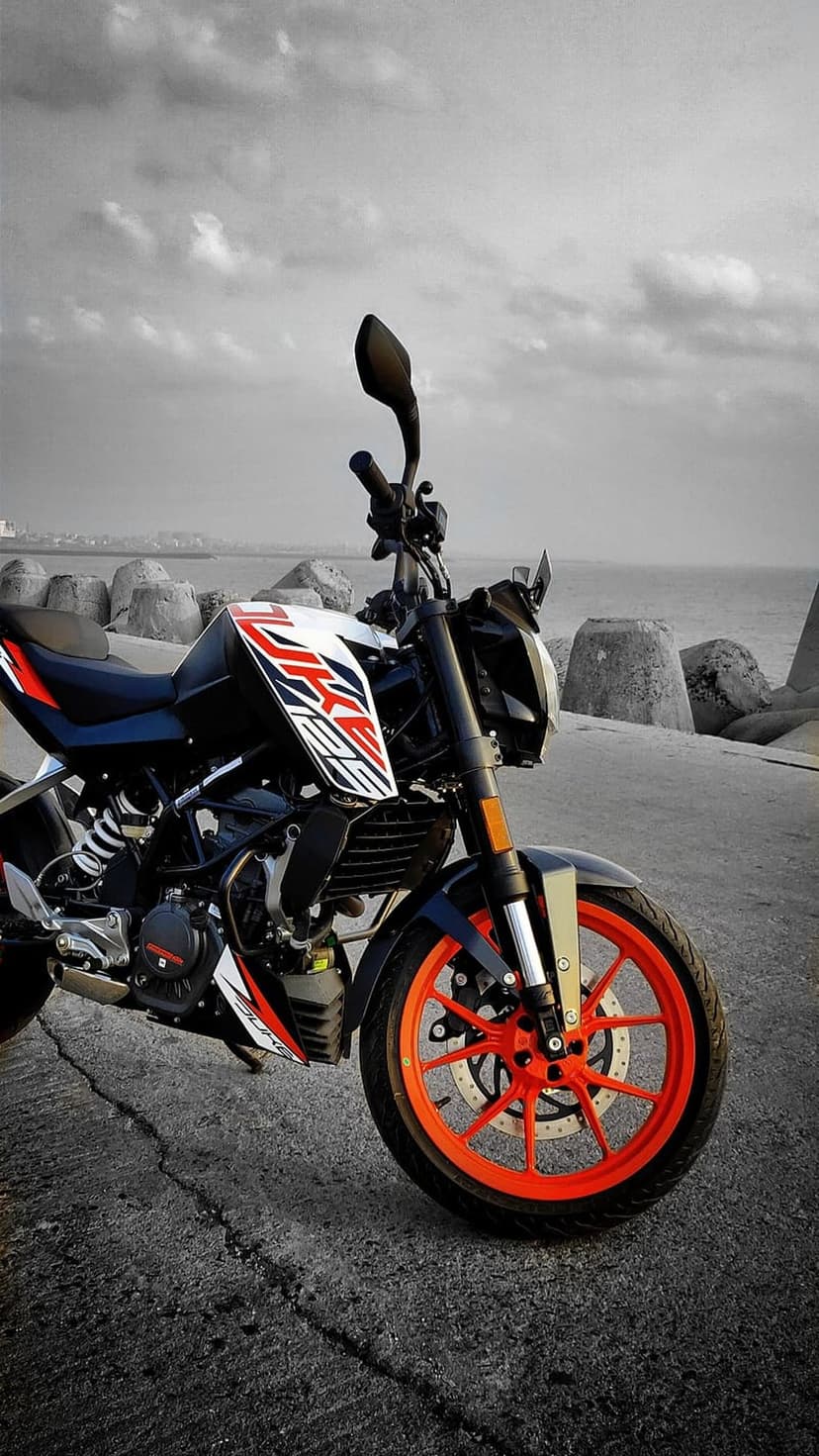 KTM Duke 125 motorcycle with orange rims parked on a cracked road by the sea under a dark and moody cloudy sky.
