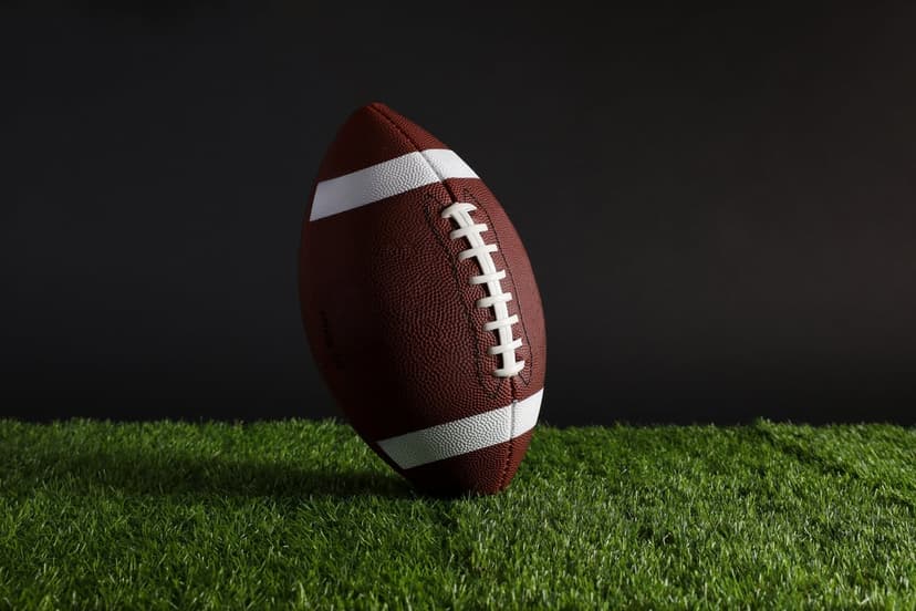 American football with white laces lies on green artificial grass against a dark, blurred stadium background.