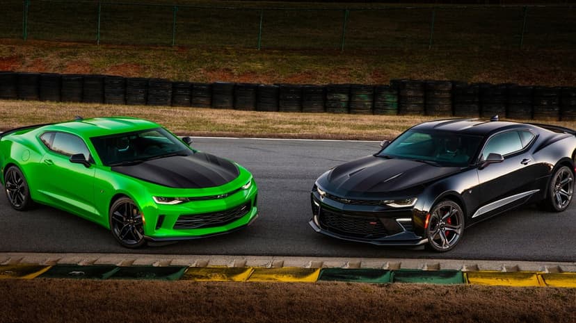 Two Chevrolet Camaro muscle cars, one bright green and one black, parked side-by-side on a paved race track.