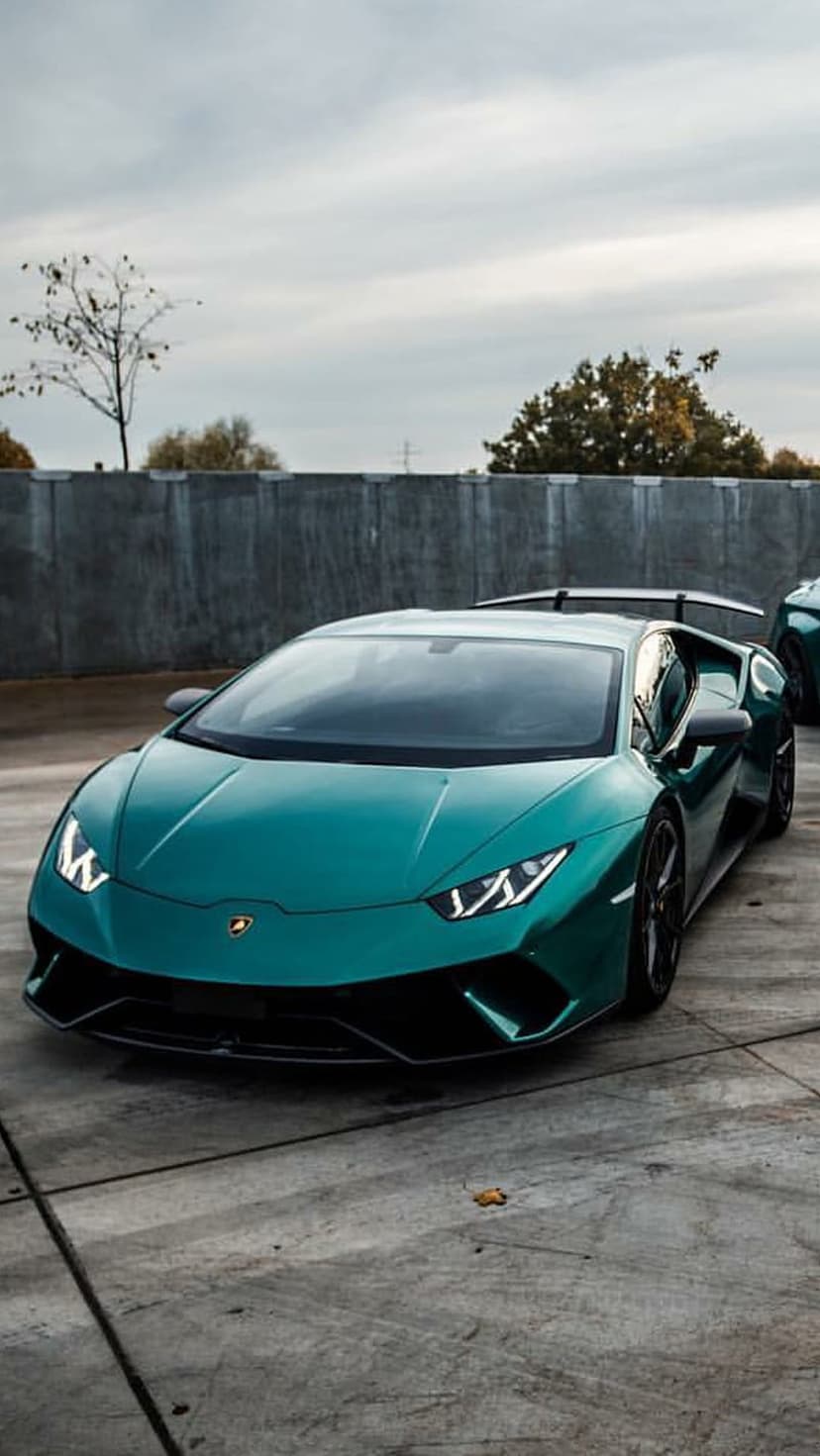 Teal Lamborghini Huracan EVO with lit LED headlights sits on a concrete lot before a grey overcast sky and trees.