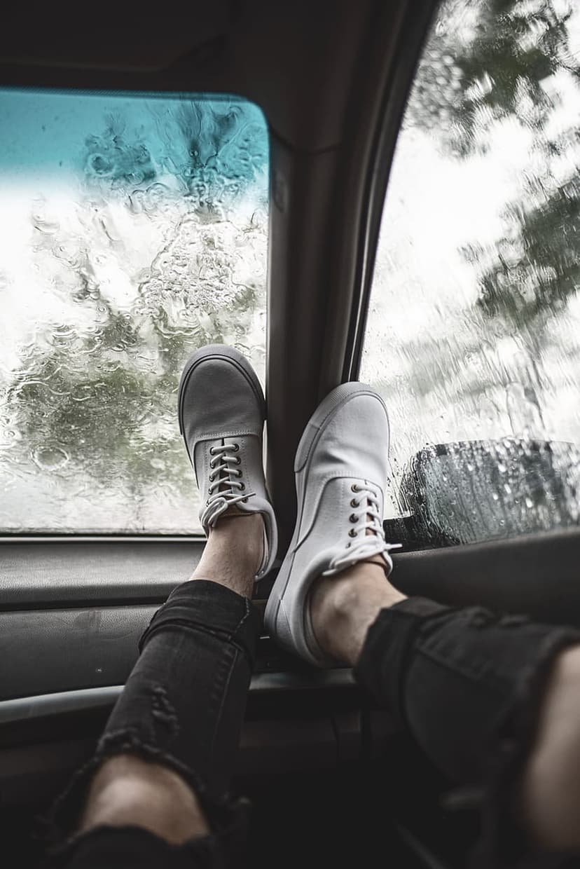 White sneakers and ripped black jeans rest on a car dashboard while rain blurs the trees visible through glass.
