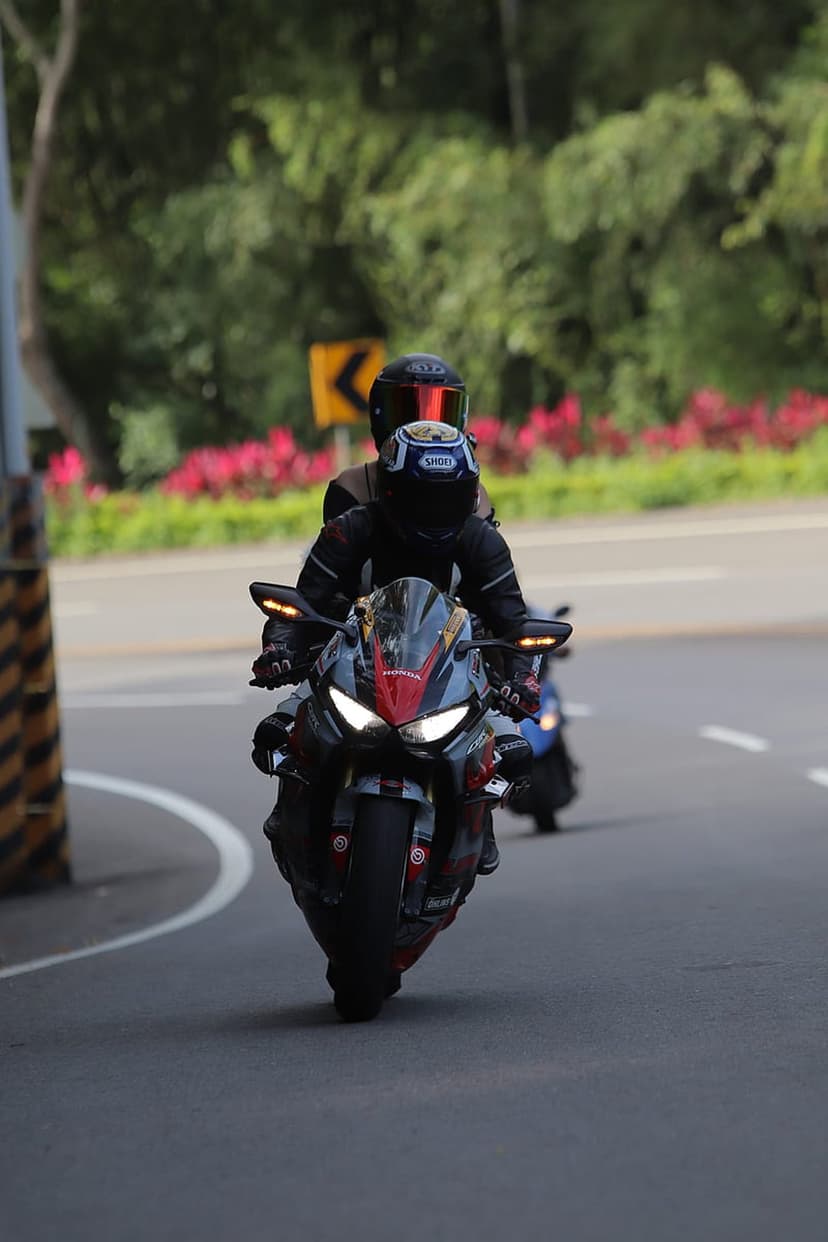 Two motorcyclists in full gear ride a red sportbike along a winding road lined with green trees and pink flowers.