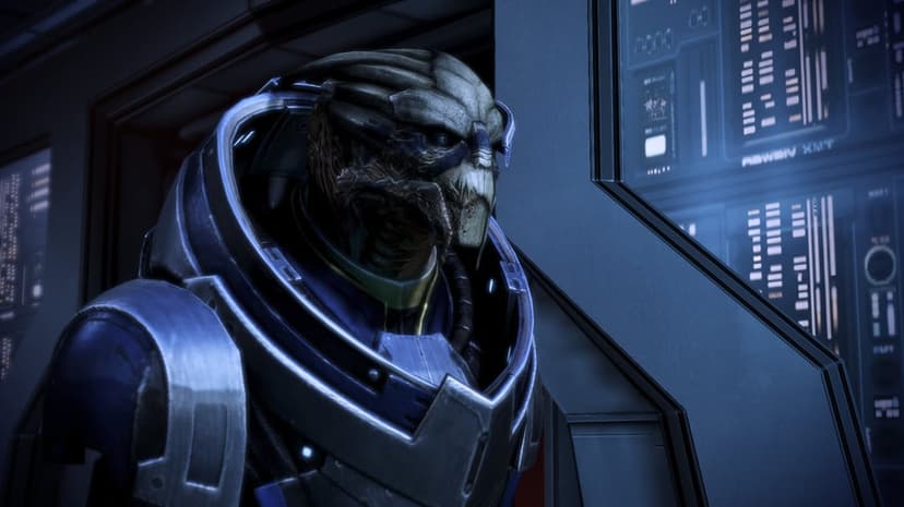 Garrus Vakarian in blue armor with facial scars stands before glowing blue computer screens in a sci-fi room.
