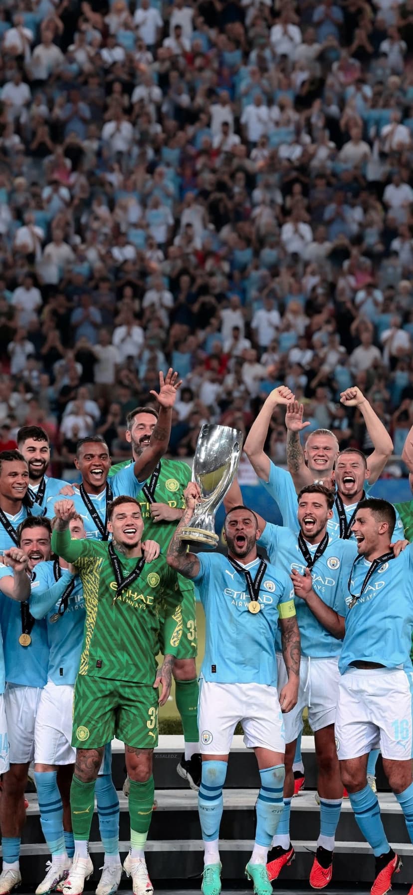 Manchester City players lift the silver UEFA Super Cup trophy overhead while cheering in a crowded stadium.