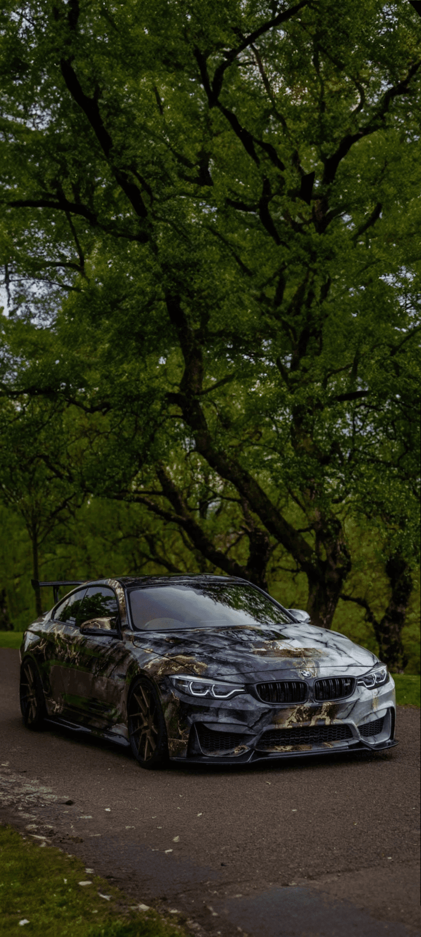 BMW M4 with a black and gold marble wrap parked on a paved forest road with dense green trees in the background.