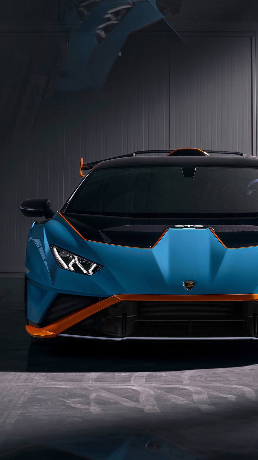 Front view of a blue Lamborghini Huracán STO with orange accents and lit LED headlights in a dark studio.