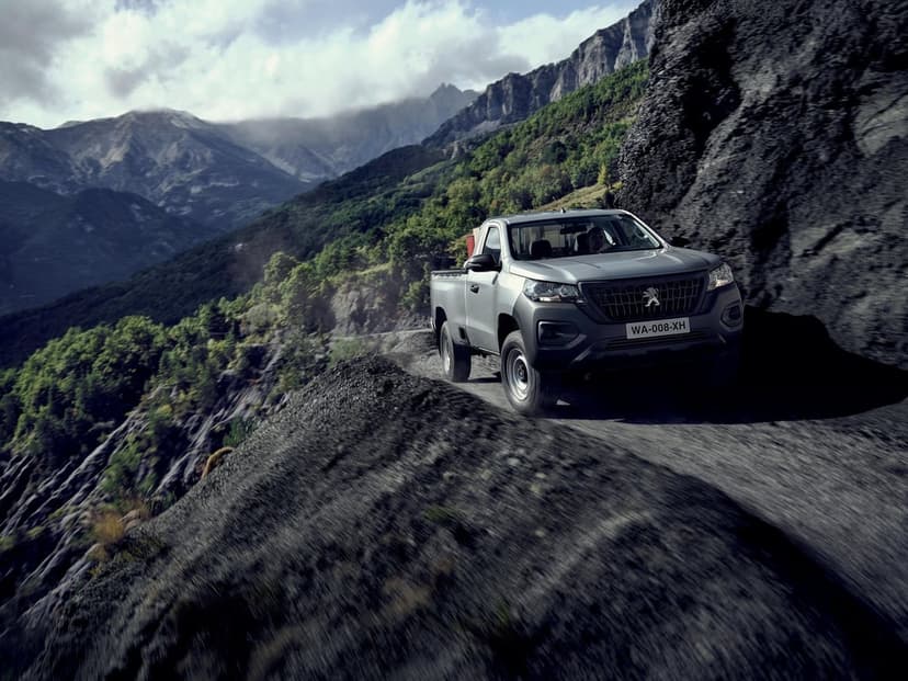Silver pickup truck driving on a narrow dirt cliff road with layers of green mountains under a cloudy sky.
