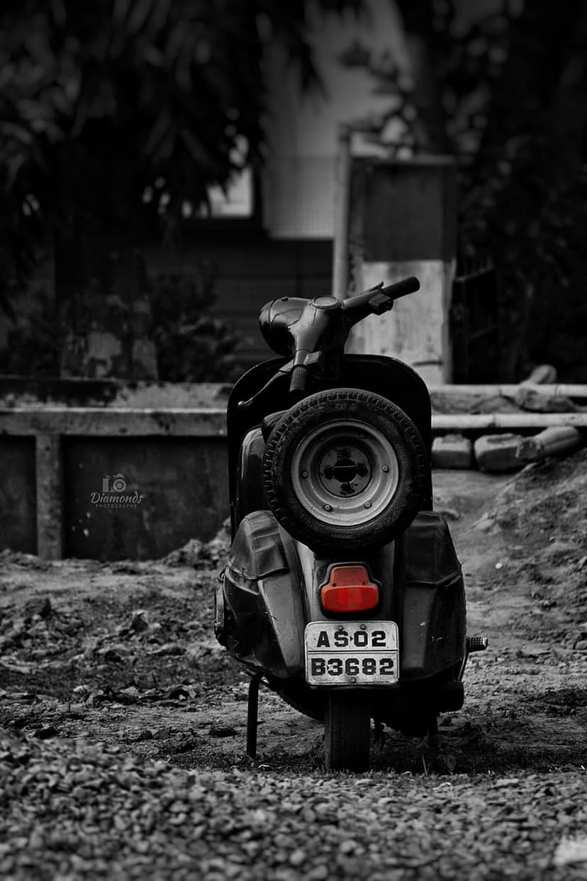 Vintage scooter with a glowing red taillight and rear spare tire parked on a grey gravel surface outdoors.