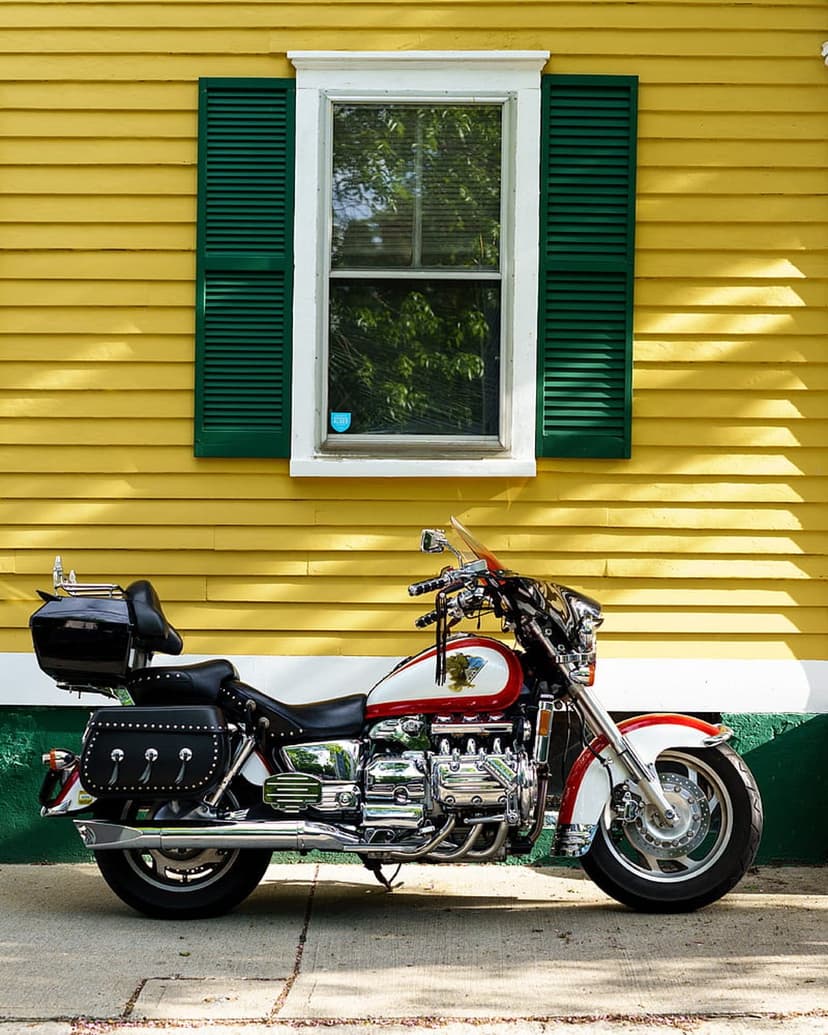 Red and white cruiser motorcycle parked in front of a yellow clapboard house with green shutters and sunlight.