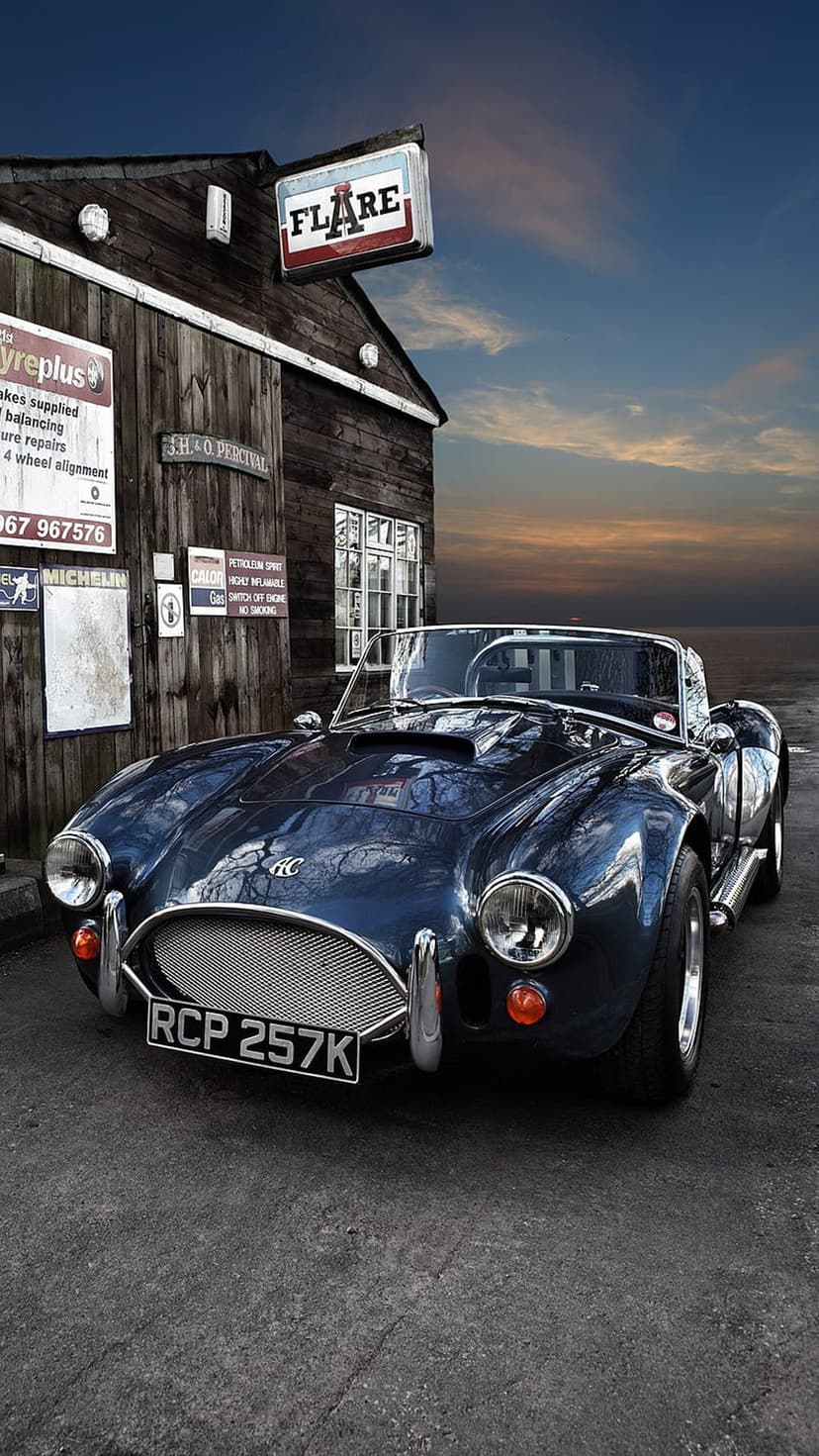 Dark blue AC Cobra roadster with chrome accents parked before a weathered wooden barn during a vibrant sunset.
