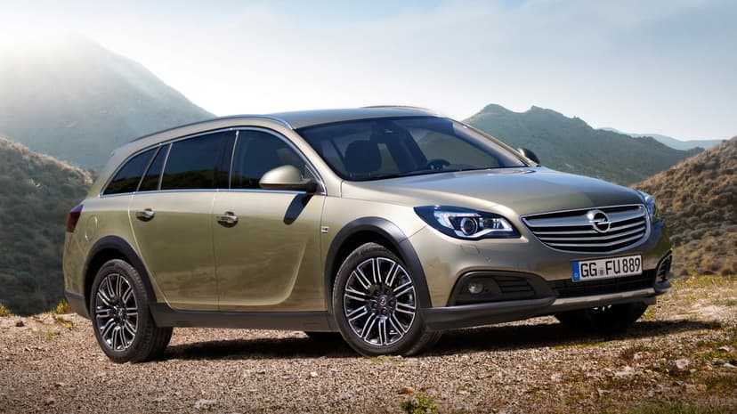 Champagne Opel Insignia Country Tourer parked on a gravel trail with sunlit mountains and blue sky behind.