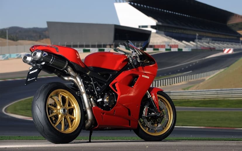 Red Ducati 1198 superbike with gold wheels parked on a racetrack with a modern grandstand in the background.