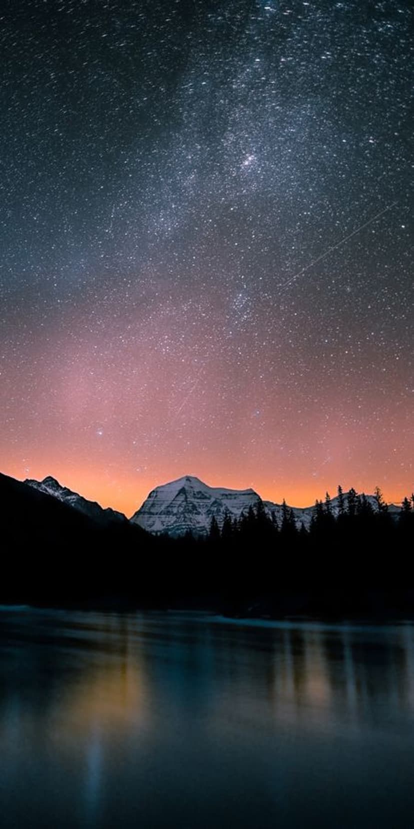 Snow-capped mountains and a starry galaxy reflect in a calm lake during a clear, crisp night in the wilderness.