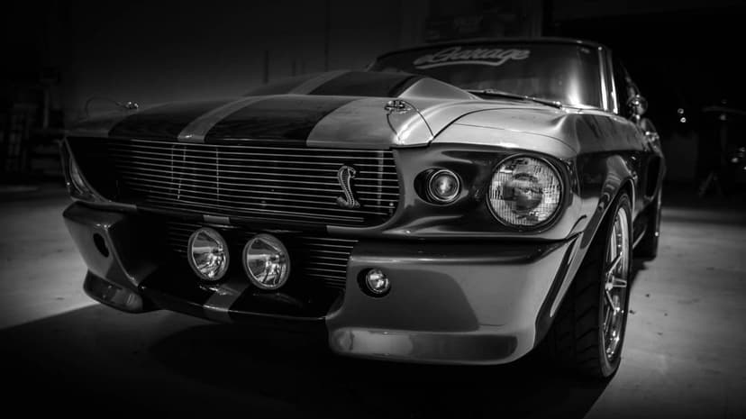 Close-up black and white front view of a classic Ford Mustang Shelby GT500 with racing stripes in a dark garage.