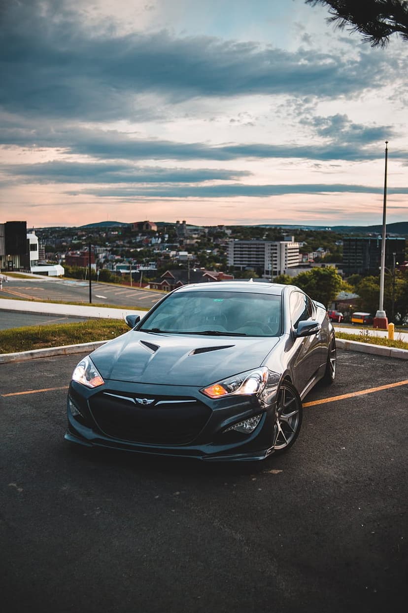 Dark gray Hyundai Genesis Coupe with headlights on parked on asphalt against a sunset cityscape and cloudy sky.