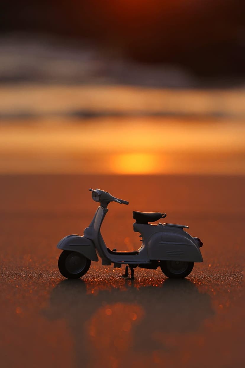Miniature blue Vespa scooter resting on wet beach sand reflects the orange and pink sunset sky during dusk.