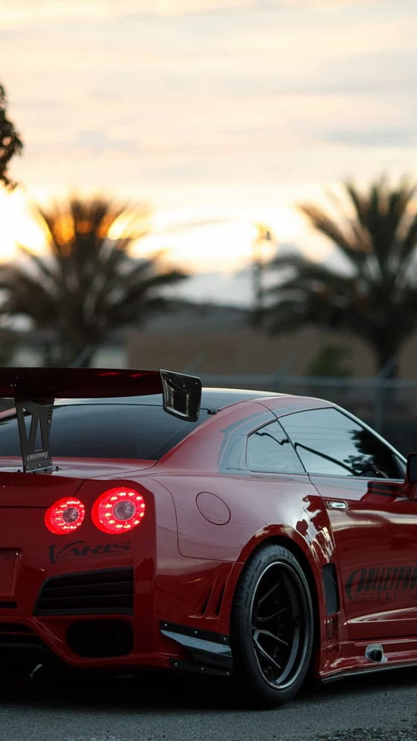 Red Nissan GT-R sports car with a large rear wing parked under palm trees during a warm golden hour sunset.