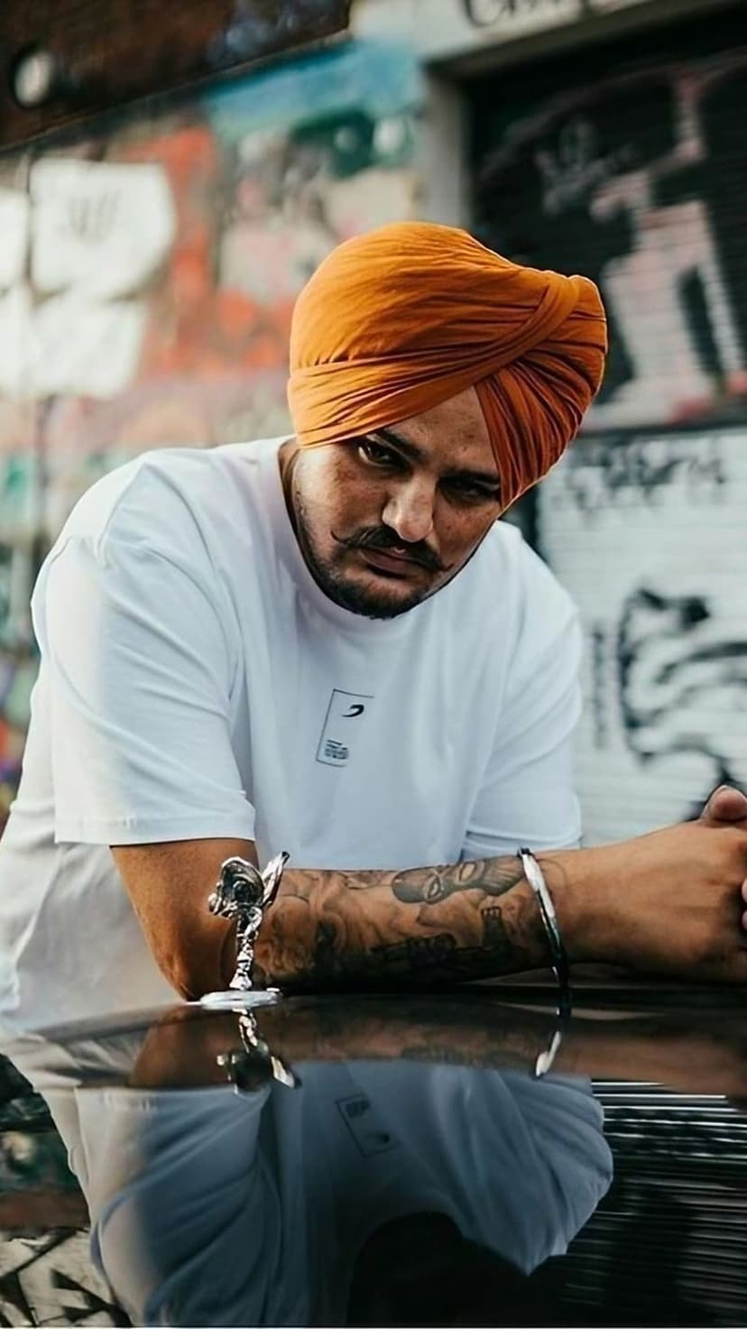 Sidhu Moose Wala wears an orange turban and white t-shirt with detailed arm tattoos and a silver bracelet.