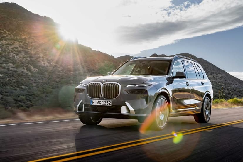 A gray BMW X7 SUV drives along a desert mountain road with a bright sun flare and blue sky in the background.