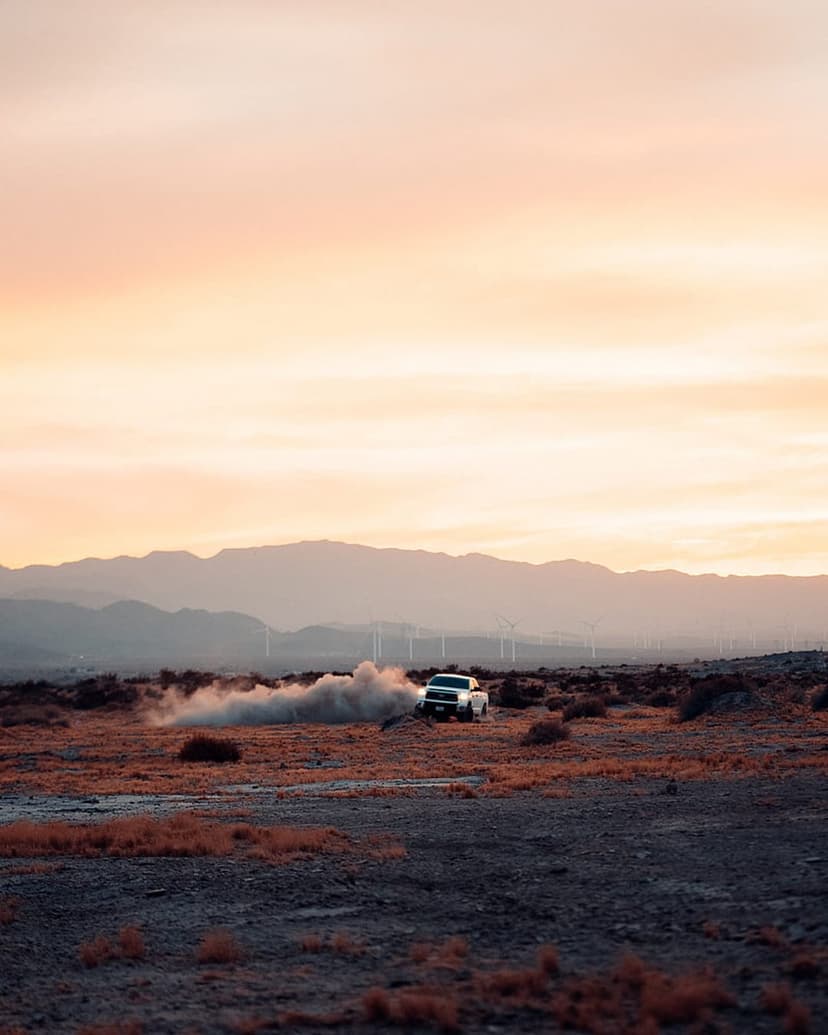 White pickup truck drifting in a dusty desert during a pink sunset with mountains and wind turbines behind.
