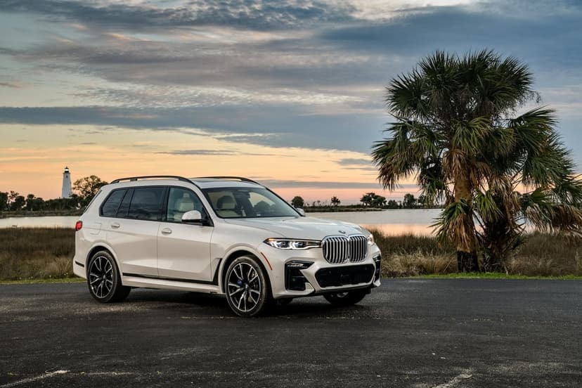 White luxury SUV parked on asphalt near a palm tree and distant lighthouse under a colorful sunset sky.