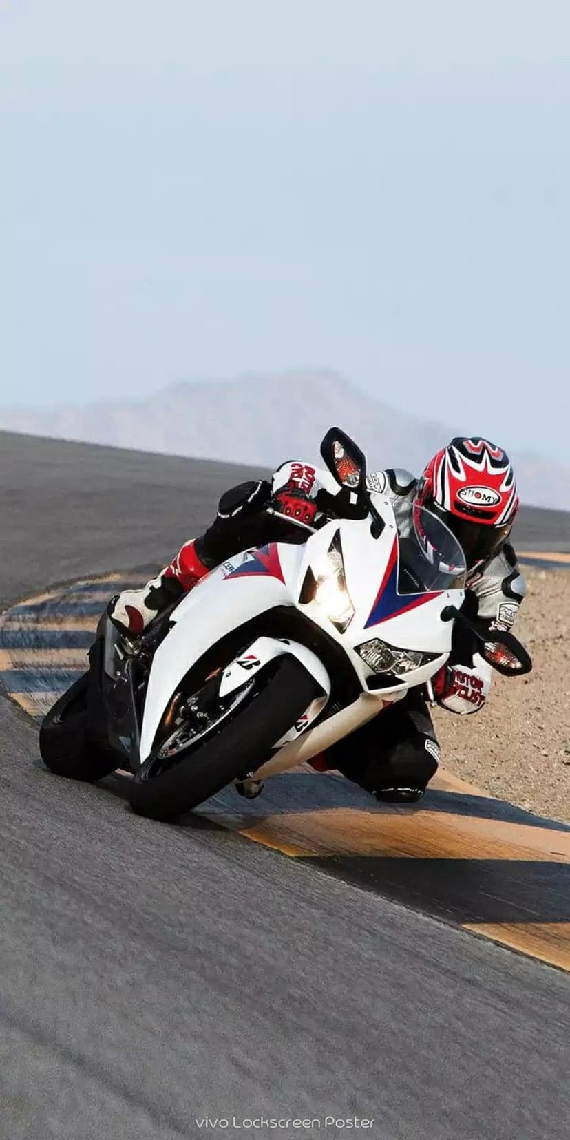 Motorcyclist in a red and white helmet leans a white sportbike into a sharp turn on a sunlit asphalt racetrack.