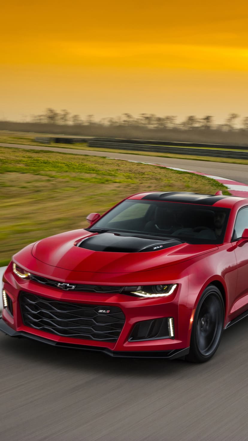 Red Chevrolet Camaro ZL1 with black hood racing on a track during a warm orange sunset with motion-blurred wheels.