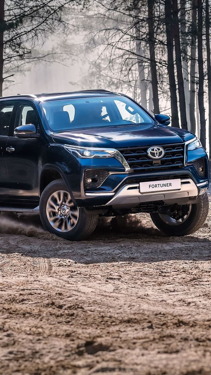Dark blue Toyota Fortuner SUV kicks up dust on a wooded dirt trail with sunlight filtering through tall trees.