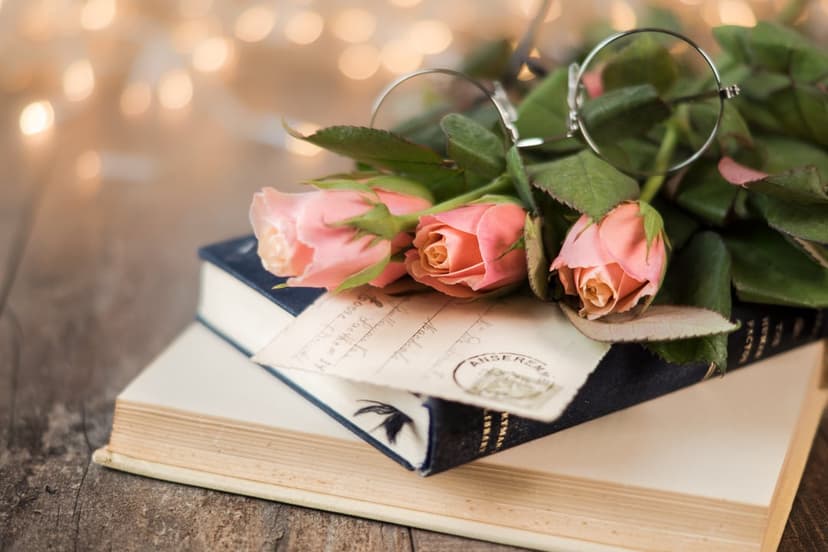 Pink roses rest on a stack of old books with wire-rimmed glasses and a sealed letter under warm bokeh lights.