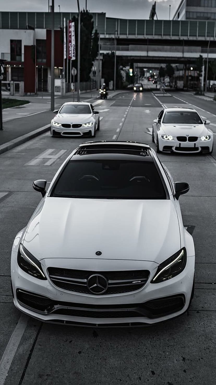 Mercedes and two BMW cars parked in a row on a city street flanked by tall buildings in bright daylight.