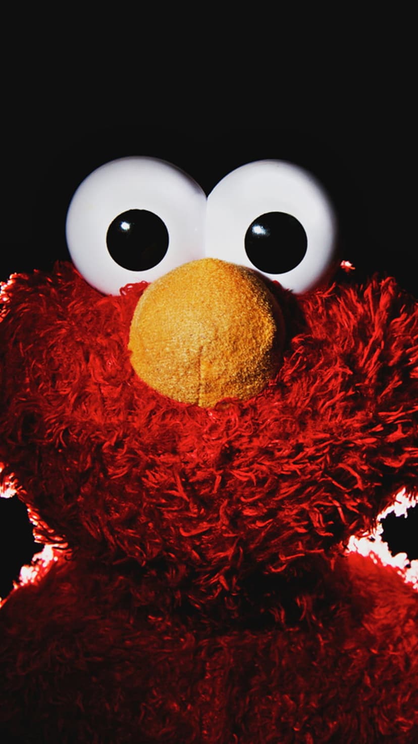 Elmo displays his shaggy red fur and large white eyes against a solid black background in a tight close-up.