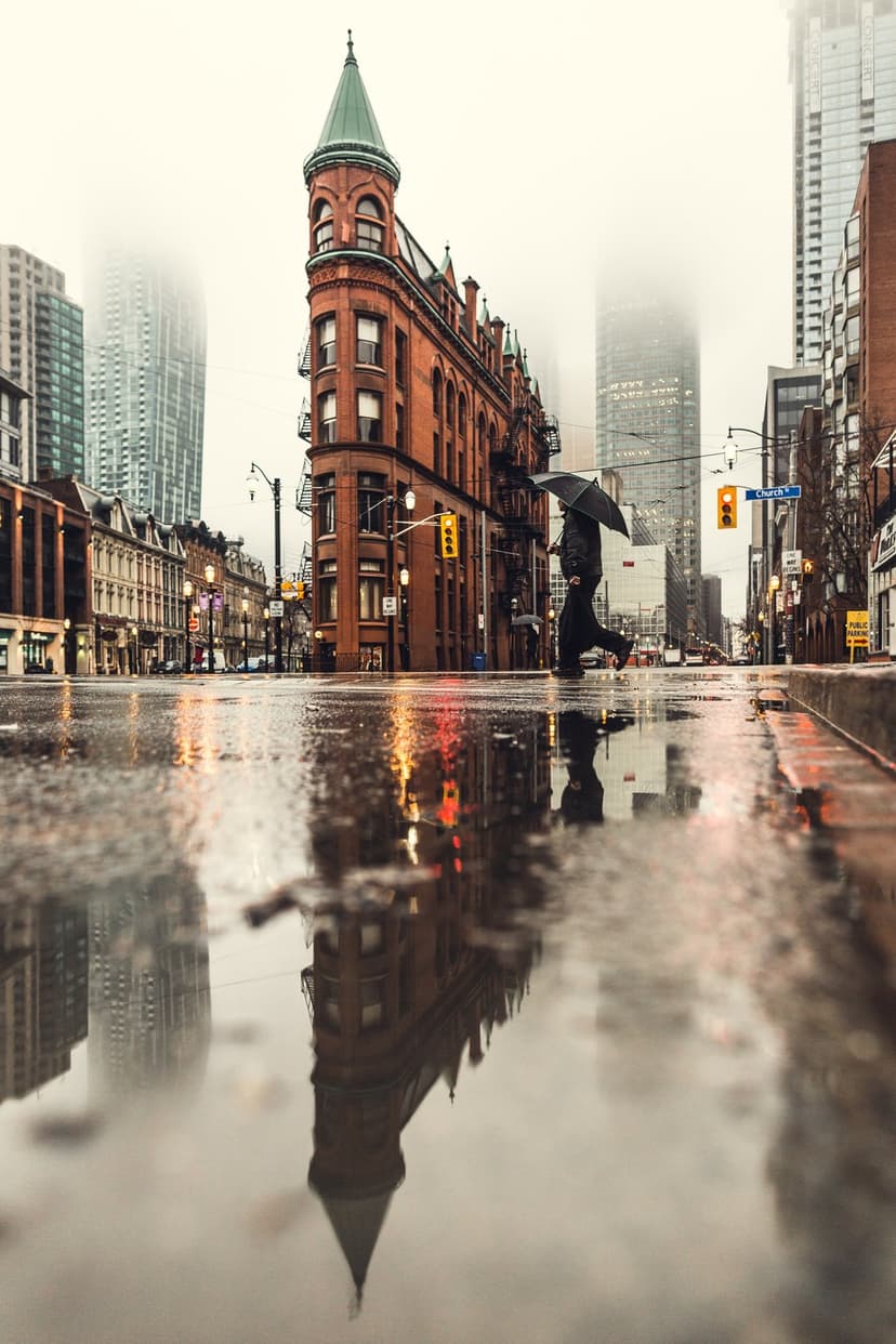 Lone figure with a dark umbrella crosses a wet city street near the Flatiron Building during a foggy evening.