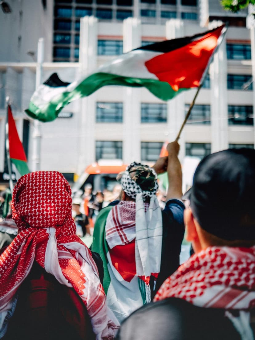 Protesters from behind wave Palestinian flags in a city street near a bus and buildings during a public rally.