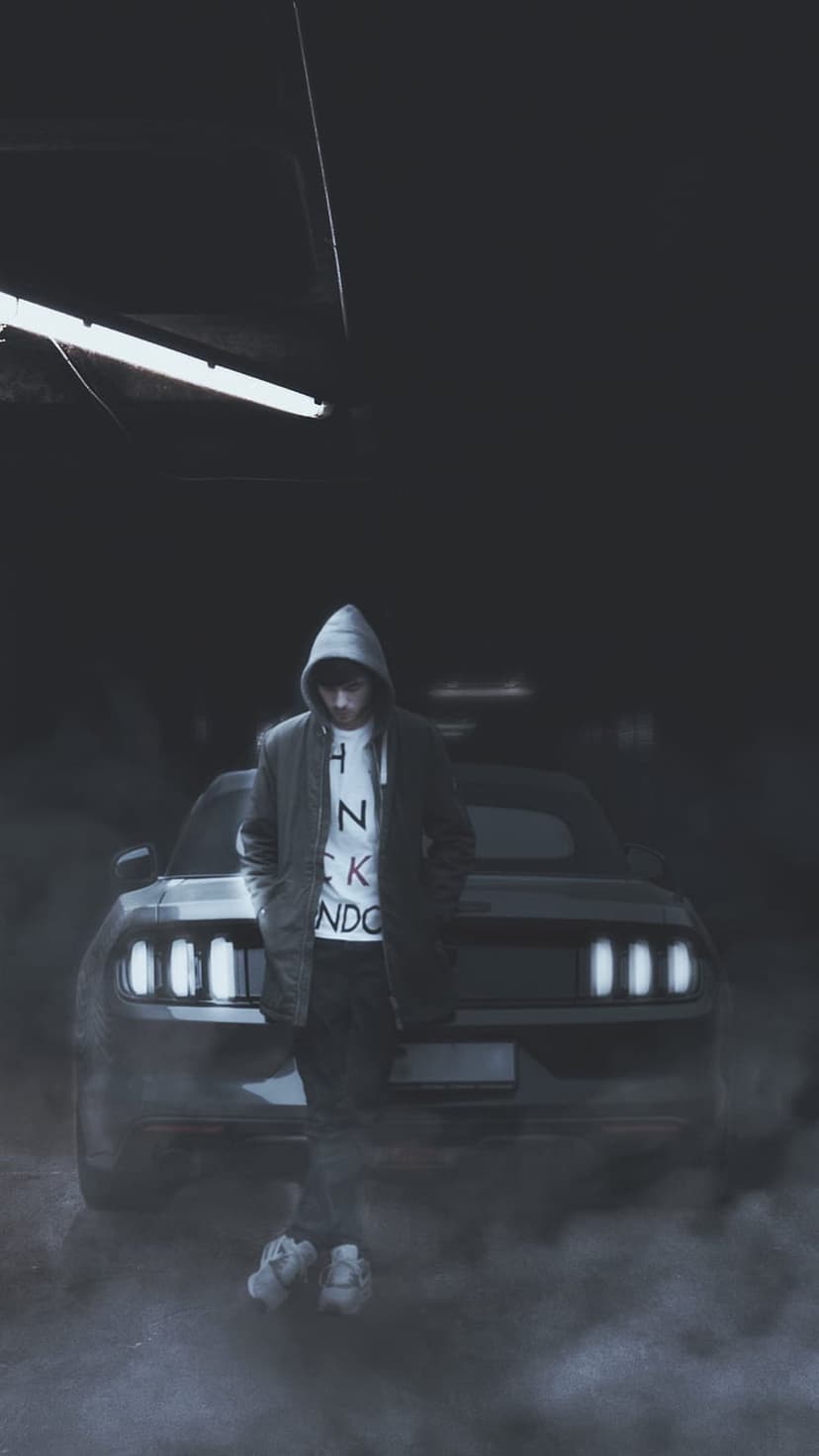 Man in a dark hoodie stands before a black Mustang with glowing headlights in a foggy, dim garage setting.