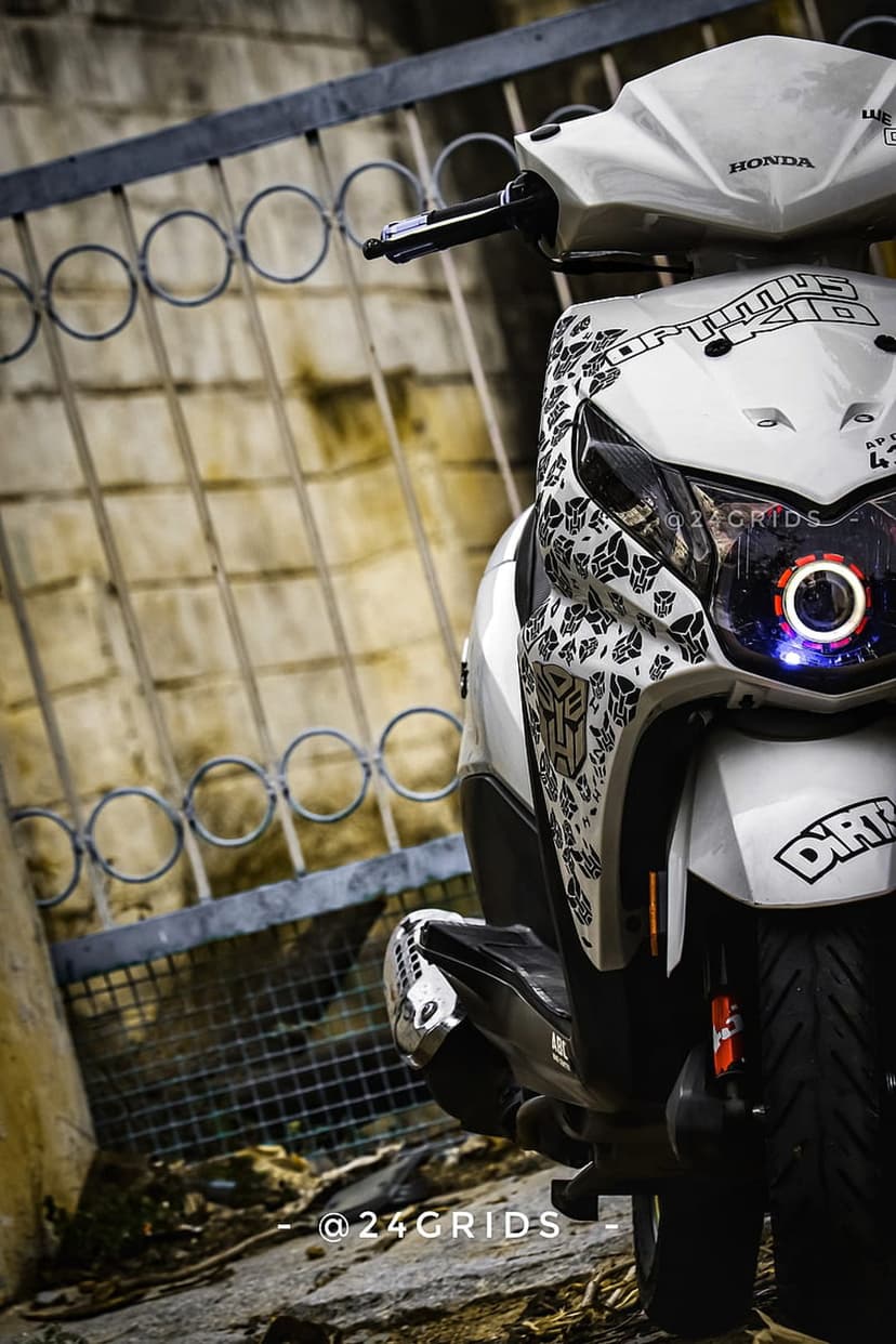 White Honda Dio scooter with black Transformers decals and a glowing LED headlight ring against a metal gate.