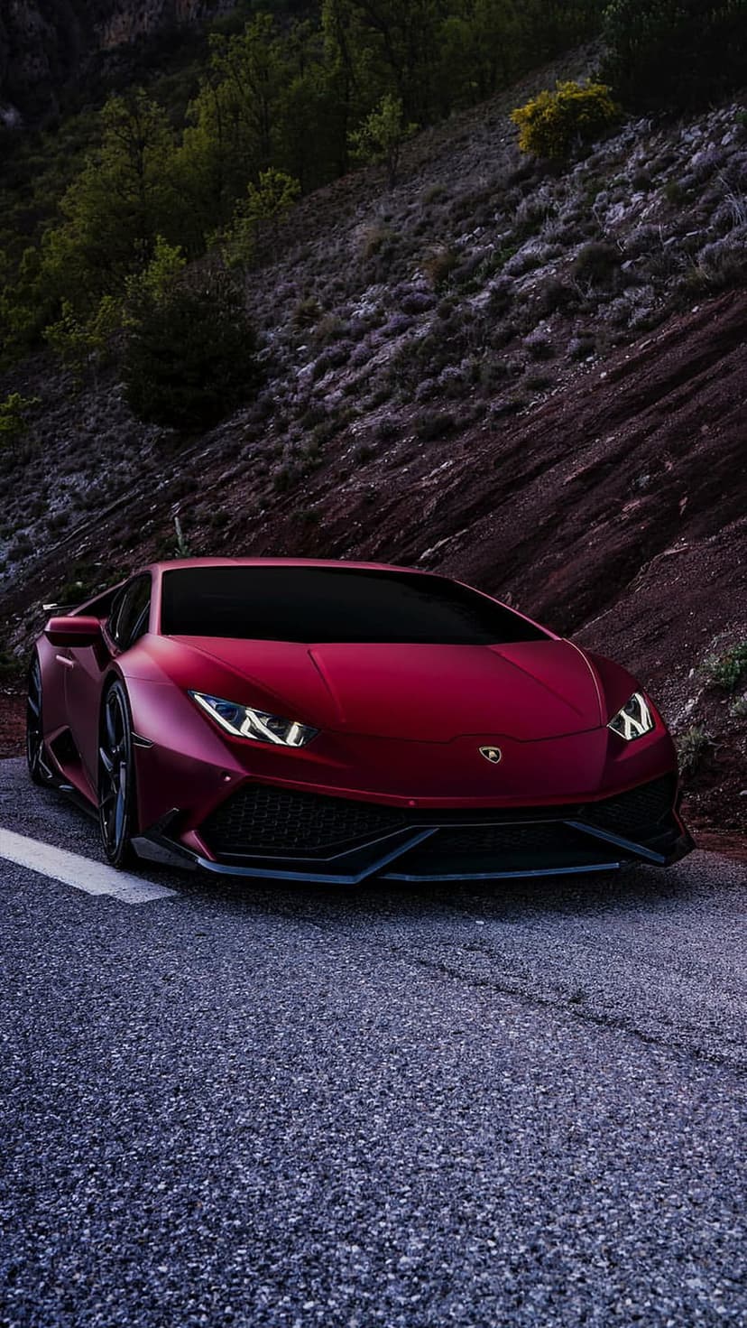 Red Lamborghini supercar on a winding mountain road with jagged rocks and green trees during golden hour light.